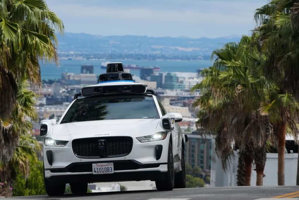 Palmiye ağaçlarıyla çevrili yokuş aşağı yolda ilerleyen tavanı sensörlü beyaz Waymo otonom Jaguar I-Pace aracı.