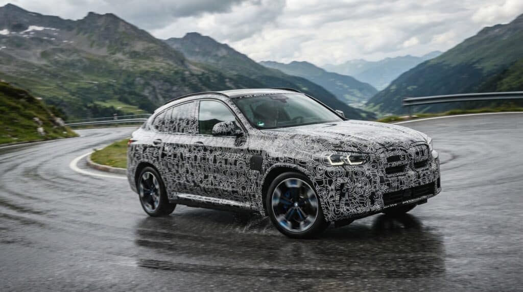 Kamufle edilmiş BMW iX4, dağ yamacındaki virajlı ıslak yolda seyrediyor.